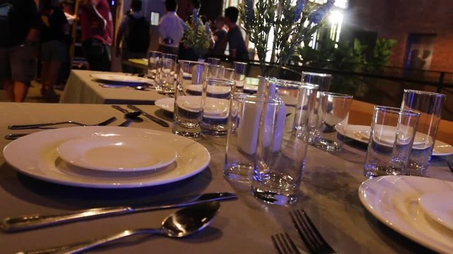 Table set with china and glassware ready for customers. Patrons of a restaurant in the background behind empty place settings. PAN RIGHT.