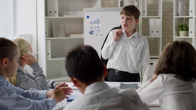 Panning of several kids sitting at desk and listening to their little business colleague walking from side to side, holding blazer and speaking