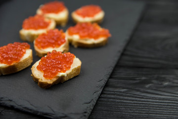  Sandwich from white baguette bread with red salmon caviar on black plate. Russian seafood cuisine. Concept of wealth and riches. 