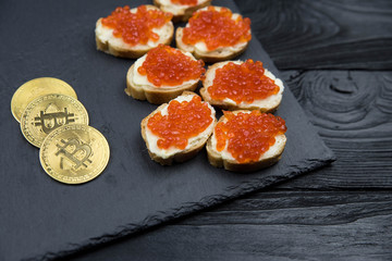 Golden Bitcoin with sandwich from white baguette bread with red salmon caviar on black plate. Concept of wealth and riches. 