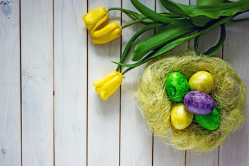 Obraz na płótnie Canvas Easter colored eggs of green yellow and purple in a basket, next to yellow tulips. Postcard for Easter, top view, there is a place for your text.