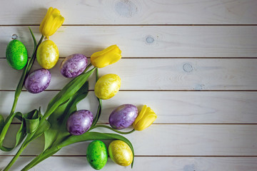 Naklejka na ściany i meble Happy easter card,copy space.Festive greeting card for Easter. On a wooden white background in the left corner a bouquet of yellow tulips and painted Easter eggs of yellow green and purple.