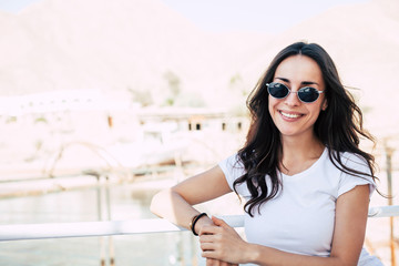 Blanc de chine. Faultless girl right in front of the camera in white t-shirt and black oval sunglasses with curly long brownish hair and sincere smile all over her hair in front of chalky buildings.