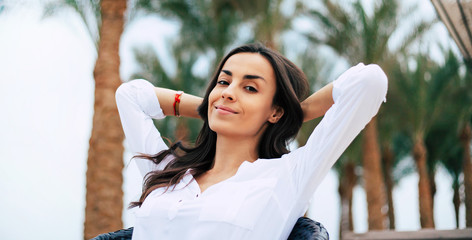 Take a breath. Cute girl in white blouse and a small accessory on her hand,  with a smiley pretty face is holding her hands aback her head and having an evening relax.
