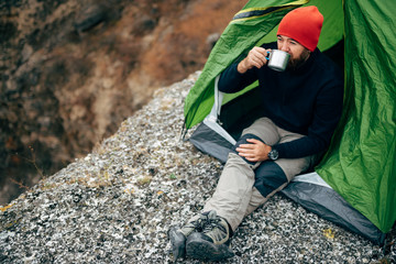 Outdoor horizontal image of young breaded man drinking hot beverage in mountains. Traveler man in red hat, sitting near to camping tent, holding in hands a mug of tea after hiking. Travel, lifestyle.