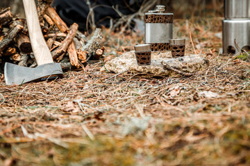 flask with glasses, camping woodstove and utensils, axe and sapper shovel near a firewoods, backpack on the background