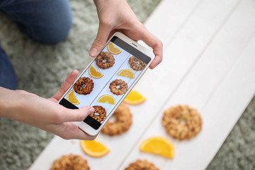 Female food photographer with mobile phone taking picture of tasty cookies and orange slices at home, closeup