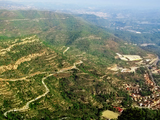 Panorama of the Montserrat mountain valley in blue fog - aerial shot, top view. Green trees, beige lines of roads and cliffs and red rectangular roofs of buildings