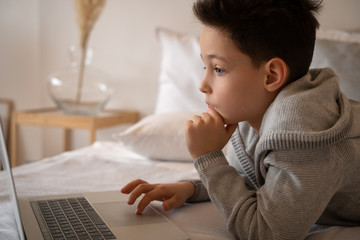 A little boy sitting on a big bed with a laptop book and looks into the camera.