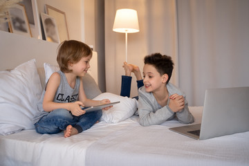 Two children, boys in parents' bed at morning with laptop and tablet. Brothers play computer games. Siblings and gadgets.