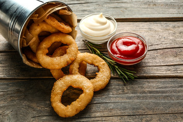 Overturned bucket with tasty onion rings and sauces on wooden table