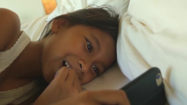 lifestyle close up head shot of sweet happy and beautiful 7 years old child having fun using internet mobile phone watching online lying on bed cheerful at night 