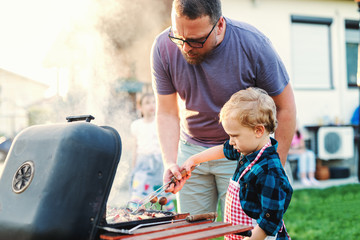 Father teaching his little son how to grill while standing in backyard at summer. Family gathering concept. - obrazy, fototapety, plakaty