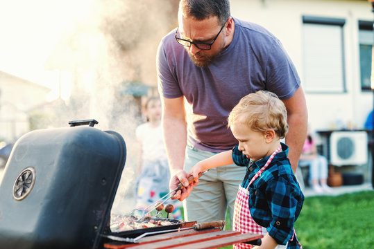 Father teaching his little son how to grill while standing in backyard at summer. Family gathering concept.