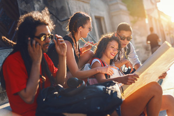 Small group of multicultural tourists sitting at steps on street and looking at map. In foreground mixed race guy talking on smart phone. Summertime.