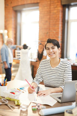 Pretty young smiling fashion designer making sketches for new collection by workplace in studio or workshop