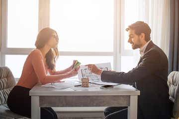 Couple buy or rent apartment together. Busy young woman hold colorful tissues in hands. Guy point on them and smile. Sit at table near window and work. Daylight.