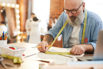 Bearded mature tailor in casualwear taking measures with tape while working over new fashion collection in studio