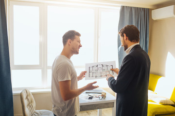 Obraz premium Young man and realtor buy or rent apartment. They hold one plan together. They talk and have business deal. Bright daylight comes out of window.