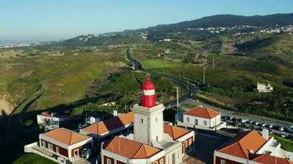Aerial; drone moves backwards from lighthouse; grassy hills, little villages of Portugal; famouse touristic sightseeing in countryside; steep slopes, rocky cliffs of Cabo da Roca; dolly zoom effect