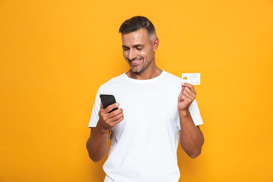 Image of beautiful guy 30s in white t-shirt holding mobile phone and credit card