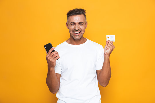 Image of handsome guy 30s in white t-shirt holding mobile phone and credit card