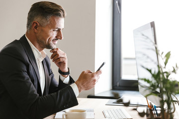 Portrait of attractive businessman using mobile phone while working on computer in office