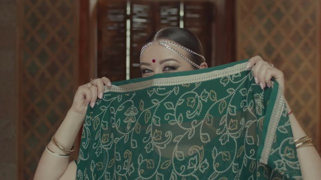 Portrait of playful female covering face with traditional indian sari, looking alluring and seductive in oriental interior. Mischievous woman in hindu dress with teasing gaze flirting indoors.