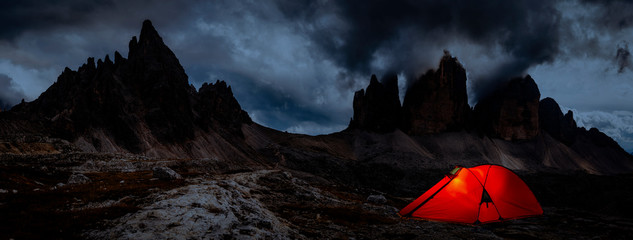 Night bivouac at Tre Cime di Lavaredo, milion star hotel under night sky, red illuminated tent on pass in Dolomites, Italy.