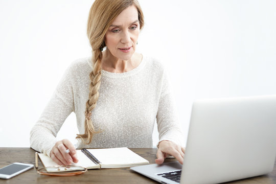 Picture of beautiful modern senior woman wearing her long braid on one side having concentrated focused look while using generic laptop and writing down in copybook, studying online at desk