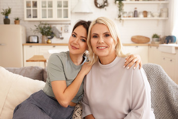 People, family and Mother's day concept. Adorable cute young brunette female embracing her mature mother, happy to spend time together, both sitting on comfortable couch in living room and smiling