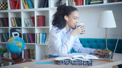 Young woman is reading up for examinations in campus library in the university. Young african american student is drinking coffee while reading articles in computer