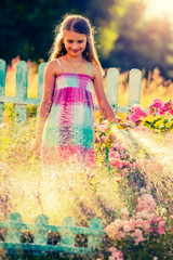 Playfull girl watering flowers with rain in the garden at summer day. Child using garden hose on sunny day at sunset lite. Little gardener playing in garden.
