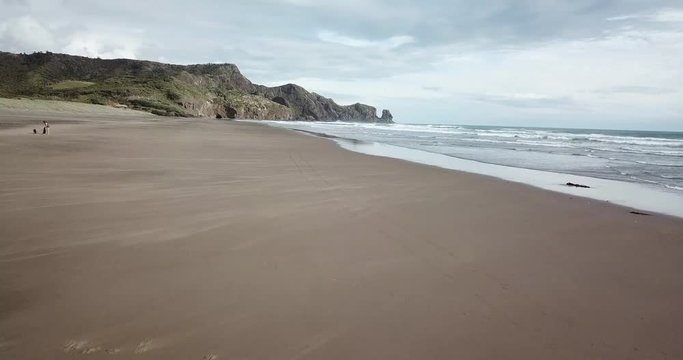 Drone shot of Bethells Beach, New Zealand with ocean waves crashing on the singular cliffs on the background, on a sunny day with some clouds