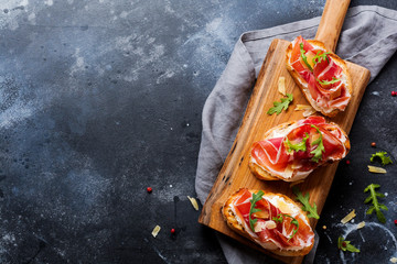 Open sandwiches with jamon, arugula and hard cheese on a wooden stand on a concrete old dark...