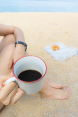 Fototapeta na wymiar Asian women sitting on beach, holding a white coffee mug with black coffee inside. Croissant is set on sand. Background is sea and sky. Concept for vacation, relaxation and tranquility.