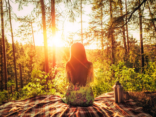 Beautiful young girl sitting on a plaid in a forest glade during sunset bright sunlight around...