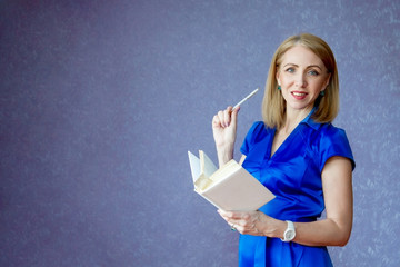 A woman in a blue dress on a purple background holding a white notebook and pen