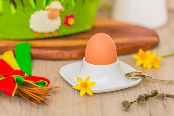 Colorful Easter decoration with hard boiled brown egg in a bowl, wooden table, spring yellow daffodil flower, braided whip made of willow, red, green and yellow ribbons, white jar and lamb decoration 