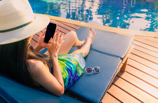 Asian woman with hat and swimsuit sit on sunbed at poolside and using smartphone on summer vacation by swimming pool. Girl with nail manicure and sunglasses. Summer vibes. Woman relaxing on holiday. - Powered by Adobe