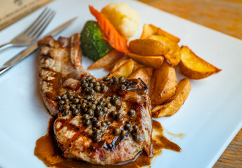 Steak topped with sauce and pepper serve with potato fried and carrots, cauliflower and broccoli steamed. Steak on white plate with stainless steel fork and knife put on wooden table. Tomahawk steak.