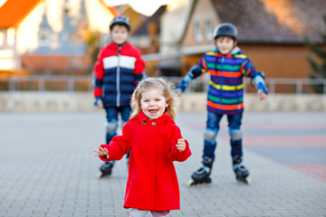 Two kids boys and little toddler girl playing together outdoors on sunny day. Brothers in protection safety clothes skating with rollers. Happy sister holding siblings. Happy family of three children