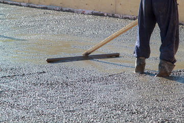 worker in rubber boots stands in uncluttered cement and leveling the surface of the base plate (fresh concrete slab) with a special wooden working tool