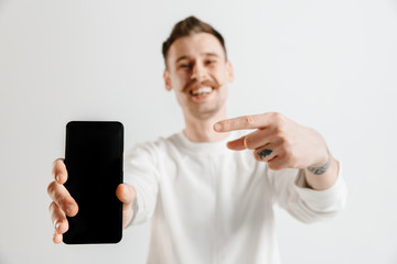 Young handsome man showing smartphone screen over gray background with a surprise face. Human emotions, facial expression concept
