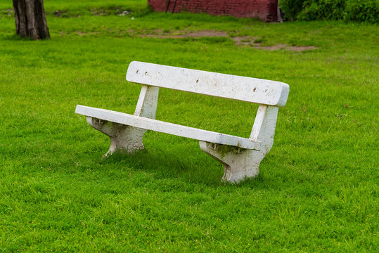 White concrete bench on a grass field with a part of a tree and a bricks construction on the back
