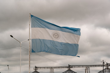 Aged Argentine flag waving in front of an industry with silos on a cloudy day