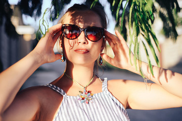 Portrait of an attractive girl in sunglasses hiding behind tree branches on a bright Sunny day, walking through the sights of a tropical city on vacation by the sea.