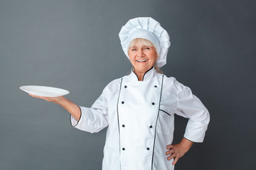Senior woman chef studio standing isolated on gray holding plate looking camera laughing cheerful