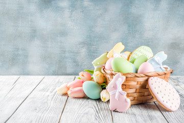Easter greeting card background with pastel colored eggs and homemade cookies shaped in eggs and bunnies rabbits. With a basket, tulips, rustic wooden table, copy space top view banner