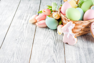 Easter greeting card background with pastel colored eggs and homemade cookies shaped in eggs and bunnies rabbits. With a basket, tulips, rustic wooden table, copy space top view banner
